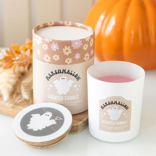 A white ghost candle in a glass jar labelled “Marshmallow Scented Candle” features a cartoon ghost; perfect for cosy autumn evenings, with its box and lid matching the design. A pumpkin and dried flowers are softly blurred in the background.