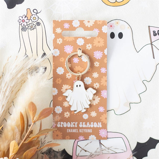 A white enamel Spooky Season Ghost Keyring on a floral card labelled “Spooky Season.” Ghost and flower illustrations on fabric evoke the Halloween spirit, with quirky seasonal accessories and dried beige plants in the corner.