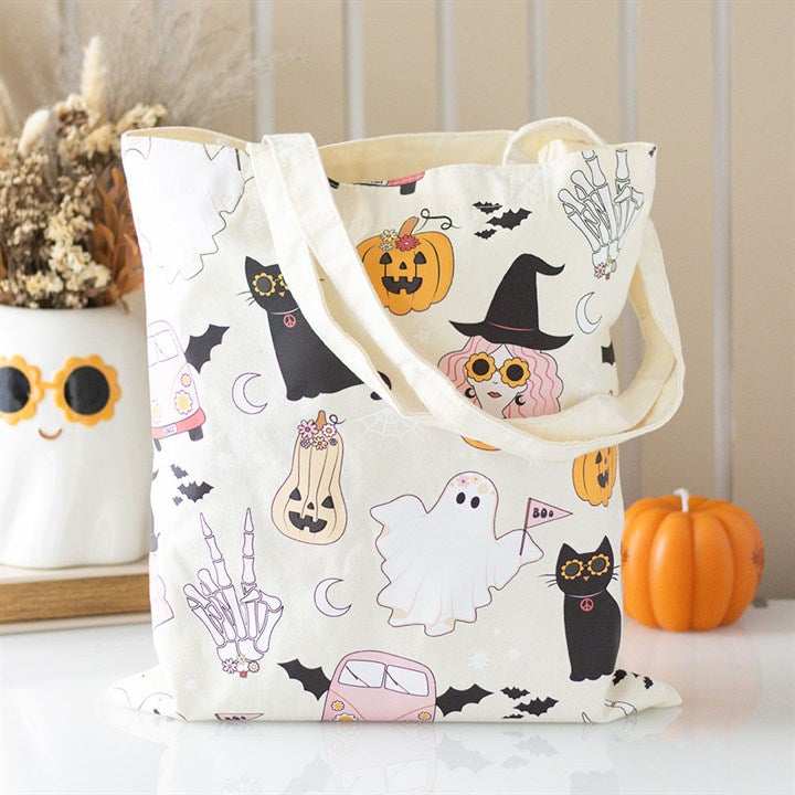 This cream-coloured Halloween tote bag displays spooky style with quirky accessories such as ghosts, black cats, pumpkins, witches, skeleton hands and bats. Shown on a light surface with festive décor in the background.