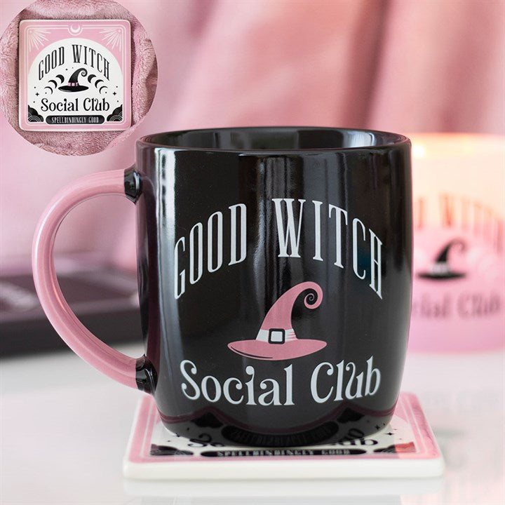 A black mug with a pink handle bears the words GOOD WITCH Social Club in white and pink lettering, enhanced by the magical touch of its witch’s hat design. The mug rests on a pink coaster, with a candle blurred in the background.