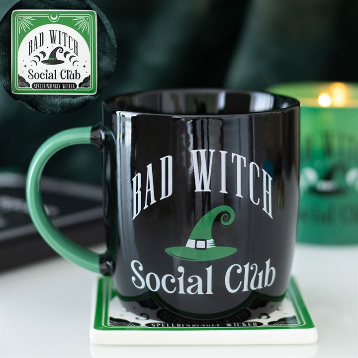A black mug with a green handle features a Bad Witch Social Club motif and a witch’s hat design, perfect for enjoying your coffee and a magical boost. It rests on a coaster, with a green candle in the blurred background.