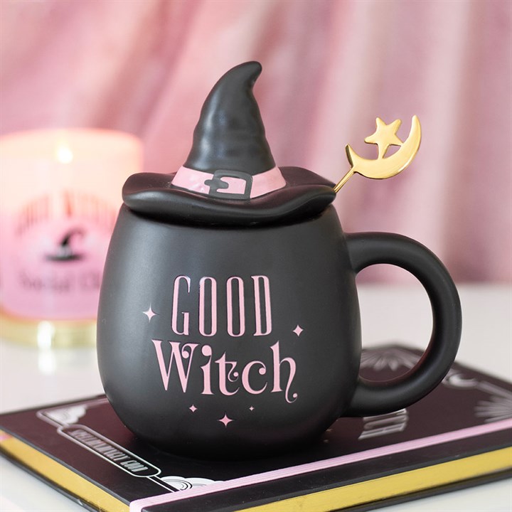 A good witch mug shaped like a cauldron with pink lettering, a ceramic lid resembling a hat, and a gold crescent moon spoon. It rests on a book, while a pink candle glows softly in the background.