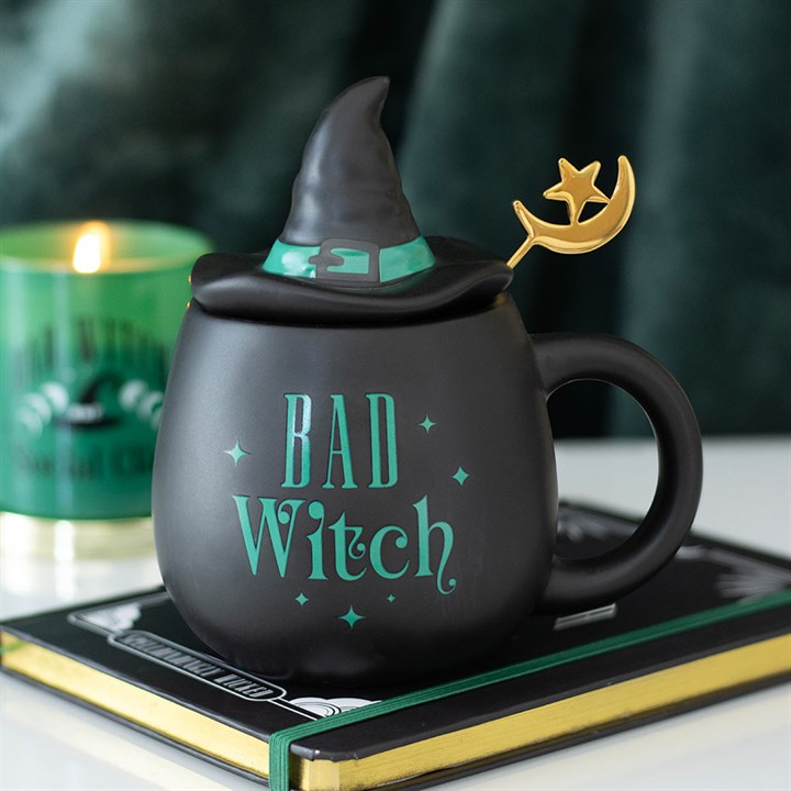 A ceramic Bad Witch mug shaped like a cauldron sits atop a book. Complete with a witch’s hat lid and a gold moon-and-star spoon, this witch hat mug exudes magical vibes beside a green candle in the background.