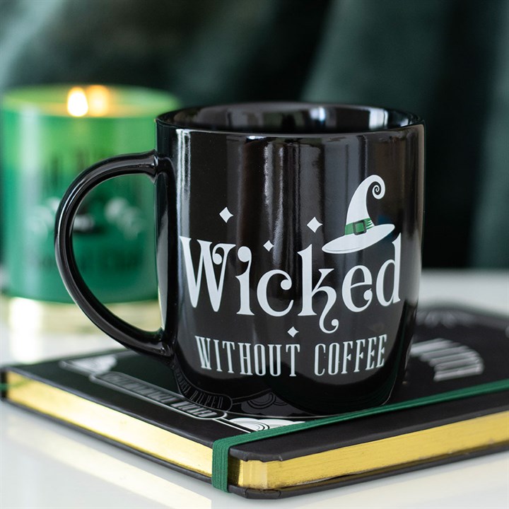 A witch’s hat mug bearing the text Wicked Without Coffee sits atop a closed book. In the background, a green candle glows, creating a cosy and magical atmosphere—perfect for your morning brews.