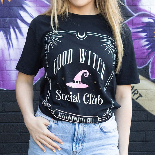 A person with long blonde hair wears a black “GOOD WITCH Social Club” T-shirt featuring a pink witch’s hat, tucked into light blue jeans—in modern witches’ style—standing in front of a purple and black graffiti wall.