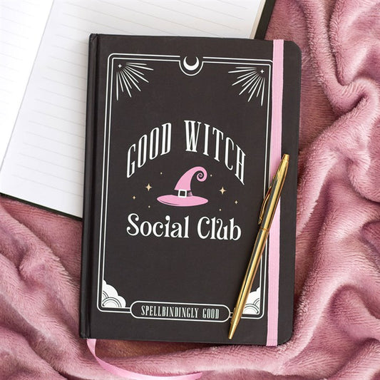 A black A5 notebook with GOOD WITCH SOCIAL CLUB and a pink witch’s hat on the cover rests on a pink blanket. A gold pen lies atop this witchy journal, with an open lined notebook partially visible beside it.