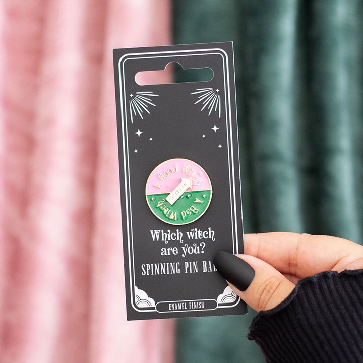 A hand with black nail varnish holds a witch accessory pin card that reads Which witch are you? The enamel spinning badge points to “Good Witch” or “Bad Witch.” Pink and green curtains appear blurred in the background.