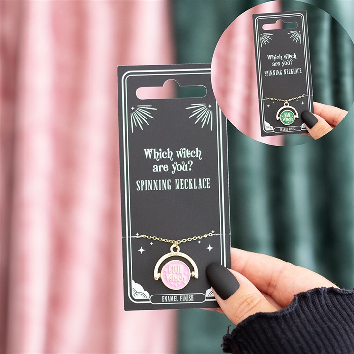A hand with black nail varnish holds a card displaying a gold spinning necklace with an enamel design—a pink and green pendant that reads “Mild Witch.” The card says “Which witch are you? Spinning necklace” in white text.