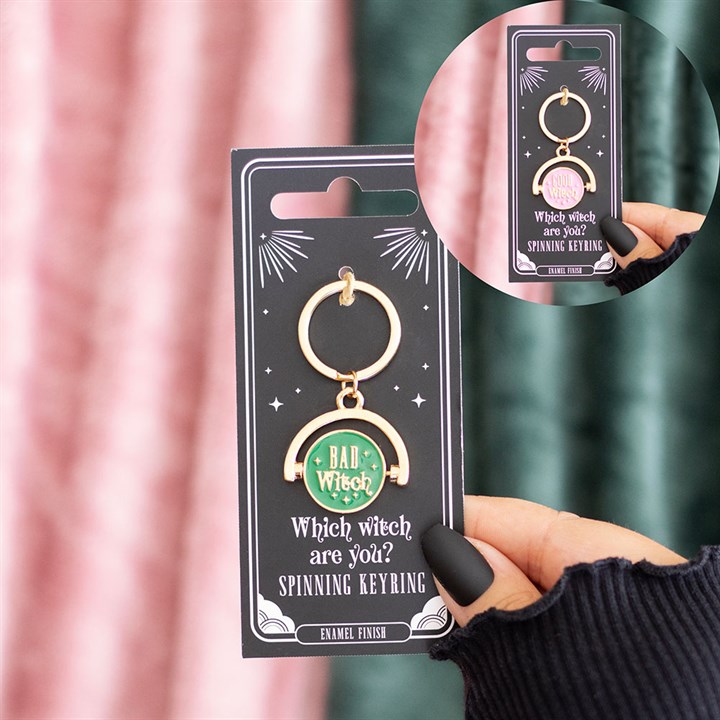 A hand with black nail varnish holds an enamel keyring on a card labelled Which witch are you? The spinning keyring reveals your witchy persona—BAD witch or GOOD witch. Curtains hang in the background.