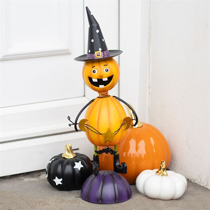 A smiling pumpkin man with a witch’s hat and pointy shoes stands on a purple pumpkin amidst orange, black (with stars), and white pumpkins—perfect Halloween decorations for autumn displays by a white door.