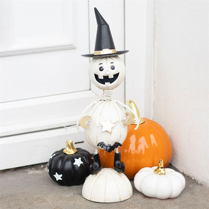 A smiling pumpkin man ornament in a witch’s hat stands by a white door, surrounded by black, orange, and white pumpkins with gold stalks—perfect for adding some spooky fun to your Hallowe’en decorations.