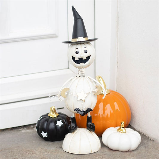 A smiling pumpkin man ornament in a witch’s hat stands by a white door, surrounded by black, orange, and white pumpkins with gold stalks—perfect for adding some spooky fun to your Hallowe’en decorations.