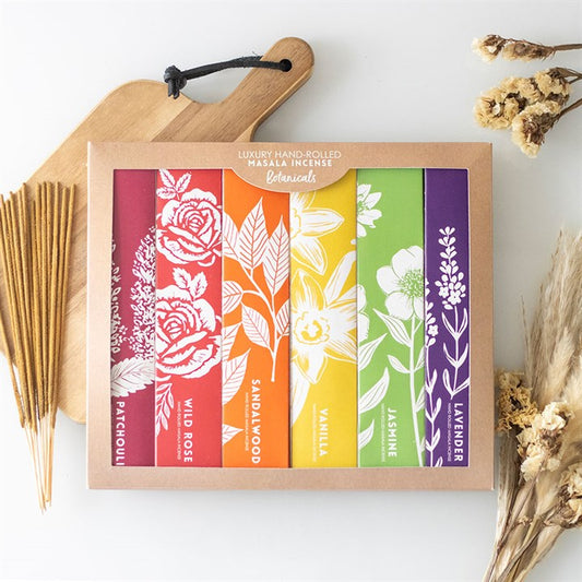 A boxed set of six hand-rolled masala incense stick packs—Patchouli, Wild Rose, Sandalwood, Vanilla, Jasmine and Lavender—with floral illustrations sits on a table alongside dried flowers and a wooden chopping board.
