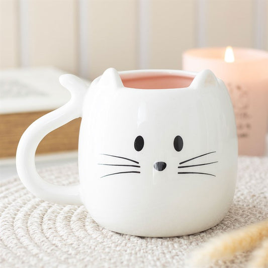 A charming cat mug, shaped like a white feline face with small ears, black whiskers, eyes, and nose. The mug rests on a woven mat, with a lit candle and a book blurred in the background.