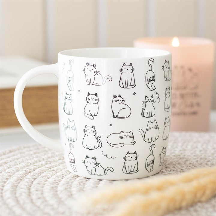 A charming coffee mug adorned with simple black line drawings of cats in various poses sits on a woven mat. A lit candle and a book are softly blurred in the background, creating a cosy atmosphere—perfect as a gift for cat lovers.