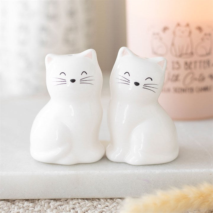 Two white ceramic cat salt and pepper shakers with simple black facial features and closed eyes sit side by side on a white surface—a charming gift for cat lovers, or a stylish addition to any cat-themed kitchen décor. A candle is blurred in the background.