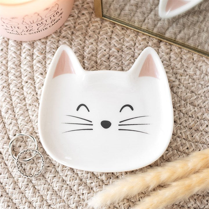 A small cat-shaped ring dish, featuring a feline face with closed eyes, whiskers, and pink ears, rests on a woven surface. Nearby are two rings, a pink candle, some dried grass, and a partially visible mirror.