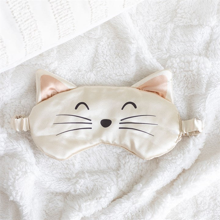 A beige satin eye mask with cat ears, an embroidered cat face, black whiskers, a nose, and closed eyes rests on a fluffy white blanket beside a textured white cushion.