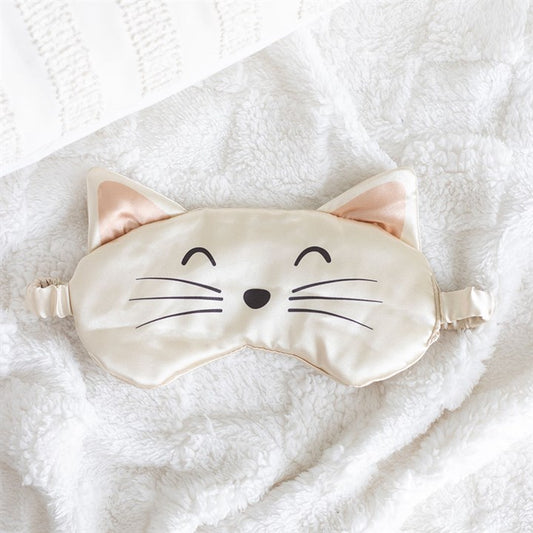 A beige satin eye mask with cat ears, an embroidered cat face, black whiskers, a nose, and closed eyes rests on a fluffy white blanket beside a textured white cushion.