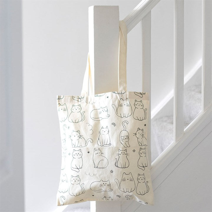A practical cat tote bag, cream-coloured with simple black line drawings of playful cats sitting and standing, hangs from a white stair bannister in a clean, bright stairwell.