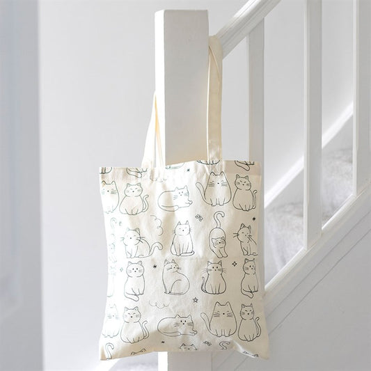 A practical cat tote bag, cream-coloured with simple black line drawings of playful cats sitting and standing, hangs from a white stair bannister in a clean, bright stairwell.