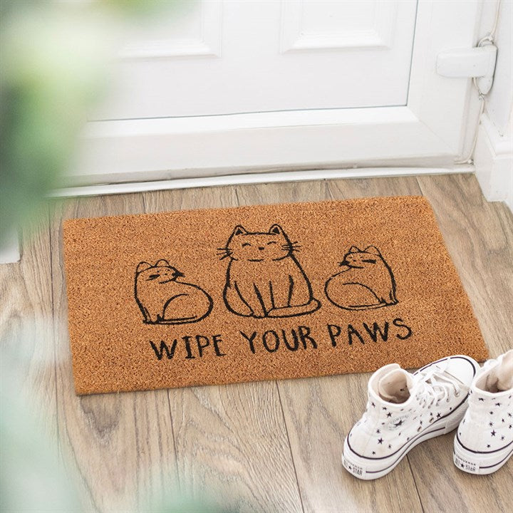 A coir cat doormat featuring three playful cats and the phrase ‘WIPE YOUR PAWS’ sits by a white door on wooden flooring, flanked by white high-top trainers and framed by blurred green leaves in the foreground.