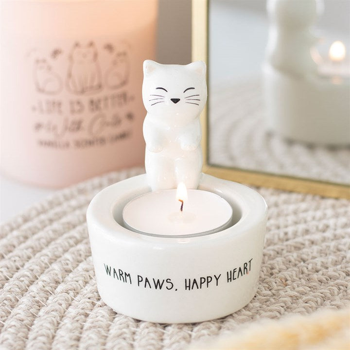 A white ceramic cat tealight holder featuring a smiling cat figure stands above a lit tealight. The holder is inscribed with “WARM PAWS. HAPPY HEART.” Soft lighting and a pink candle complete this charming gift for cat lovers and home décor.