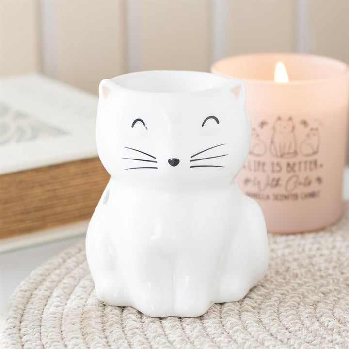 A white ceramic mug shaped like a cat, with a cute face, sits on a woven mat. Behind it, a lit candle in a pink holder decorated with cat illustrations and text resembles an adorable cat oil burner. The background is softly blurred.