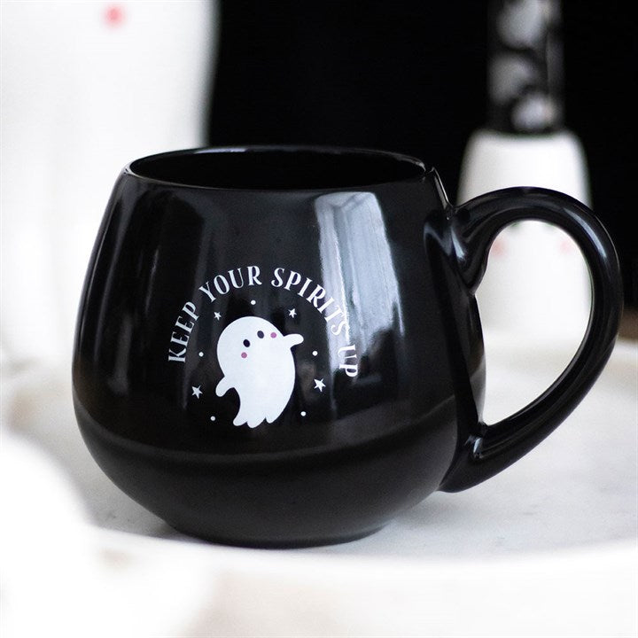 A round black Halloween mug with a handle features a cute white cartoon ghost design and the words KEEP YOUR SPIRITS UP. The mug rests on a light-coloured surface with a blurred background.