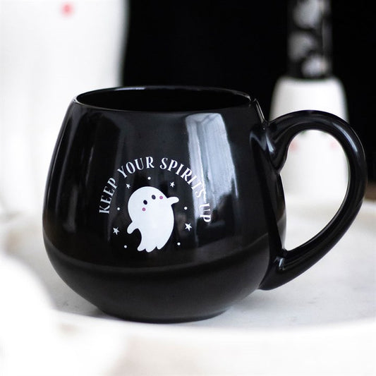 A round black Halloween mug with a handle features a cute white cartoon ghost design and the words KEEP YOUR SPIRITS UP. The mug rests on a light-coloured surface with a blurred background.