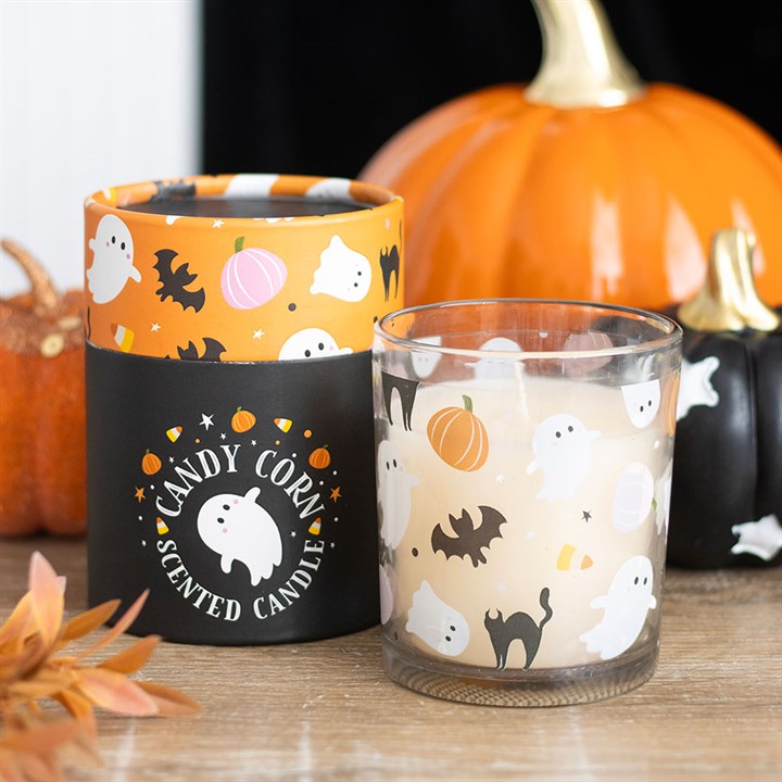 A glass and candle box featuring a ghost design and other Halloween decorations—white ghosts, bats, black cats, pumpkins, and candy corn—are arranged in front of orange pumpkins. The label reads: Candy Corn Scented Candle.