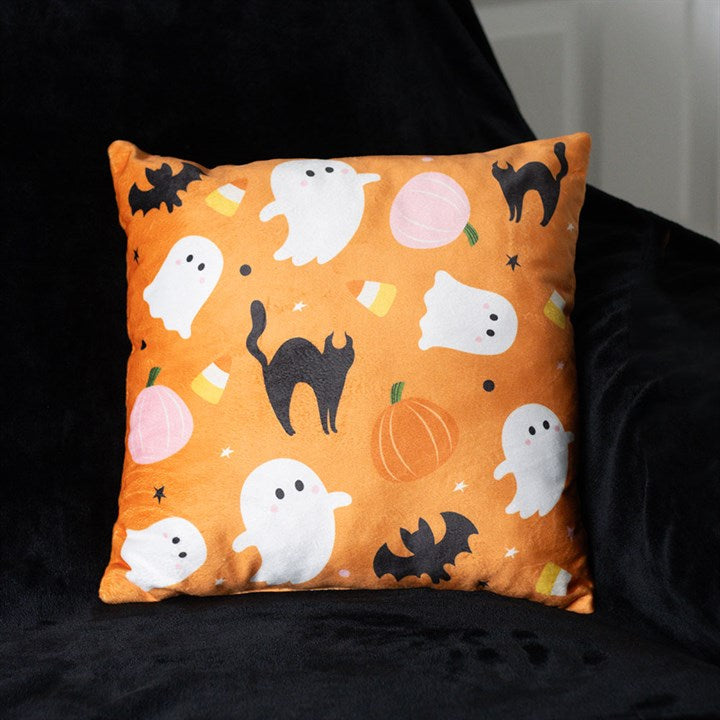 A square orange Halloween cushion featuring a spooky pattern of white ghosts, black cats, bats, pumpkins, candy corn, and stars—perfect for bringing festive charm to your autumn evenings. The cushion rests on a black fabric background.