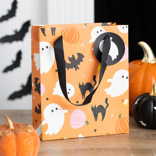 A festive orange paper gift bag decorated with white ghosts, black cats, bats, pumpkins, and sweetcorn sits on a table. Perfect for Halloween treats during the spooky season, it features black ribbon handles with bat decorations in the background.