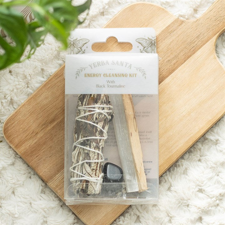 A packaged Yerba Santa Energy Cleansing smudge kit rests on a wooden board. The kit includes a sage bundle, palo santo stick, black tourmaline stone, and is partially covered by a green plant for purification.
