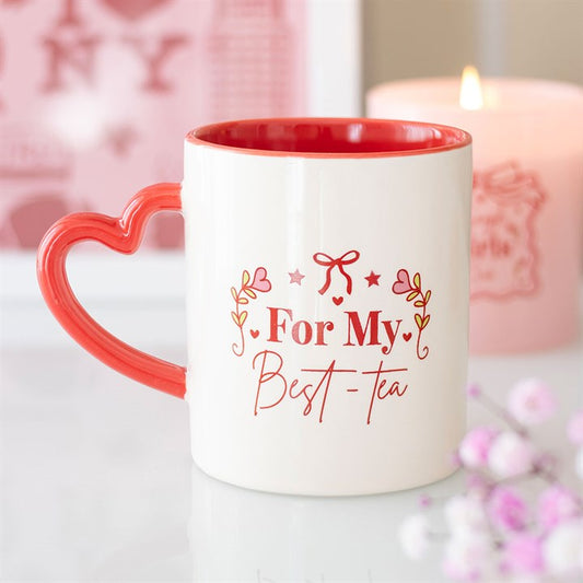 A ceramic mug with a heart-shaped handle, red interior, and “For My Best-Tea” in decorative red script—ideal as a gift. A pink candle and blurred pink flowers enhance the cosy atmosphere.