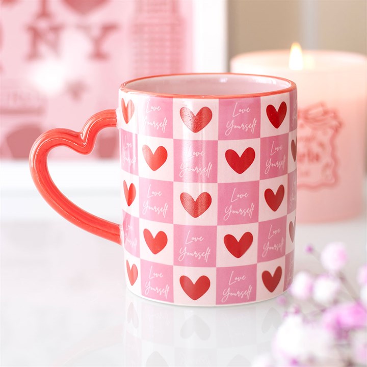 A pink and white chequered mug with red hearts and Love Yourself lettering sits on a white surface. This Love Yourself mug features a heart-shaped handle. A pink candle and blurred décor are in the background.