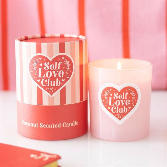 A pink Self Love Club candle with a coconut scent glows next to its matching striped box, both adorned with pink and red hearts—a lovely addition of self-care for your space.
