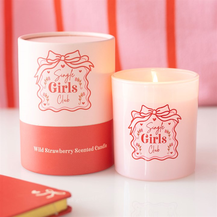 A pink Single Girls Club candle with floral designs is lit beside its matching cylindrical box, labelled “Wild Strawberry Scented Candle.” Both display red text and graphics. A red notebook sits nearby—ideal for a Galentine’s Day gift.