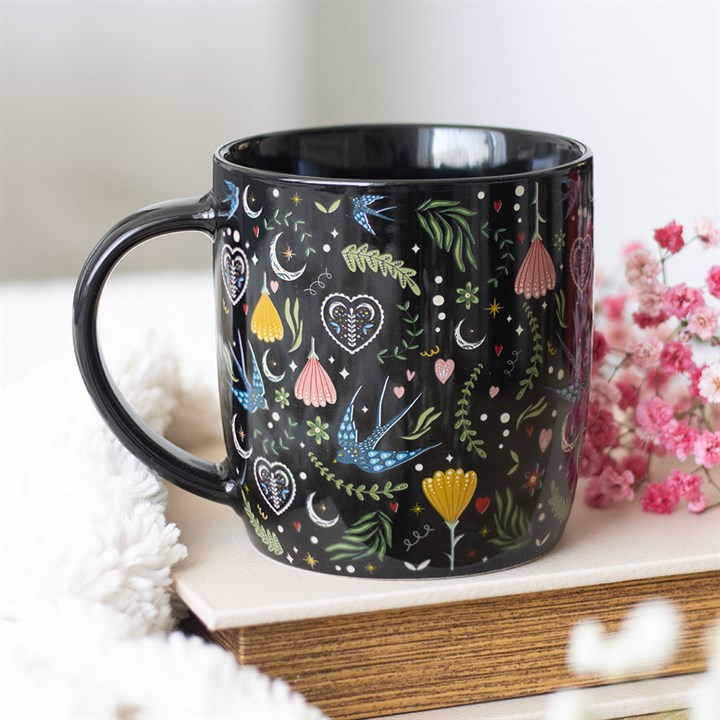 A stylish floral-print mug crafted from black ceramic, adorned with whimsical pastel botanicals, rests atop a stack of books beside pink flowers and fluffy white fabric. The softly blurred background adds charm.
