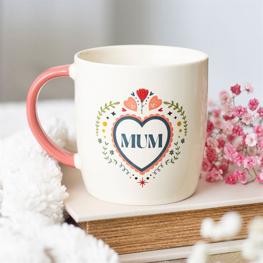 A cream-coloured ceramic mug with a pink handle rests on a book, displaying a heart motif with “MUM” and floral details—an ideal, heartfelt Mother’s Day present in this warm, inviting setting.