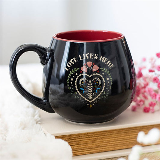 A ceramic mug with a red interior rests on a closed book. The mug displays a colourful folk heart design and floral motif, with the words LOVE LIVES HERE above it. Out-of-focus pink flowers appear in the background.