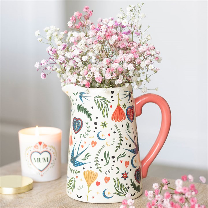 A ceramic jug with a folk floral pattern and pink handle holds babys breath flowers. Next to it, a lit candle with a heart-shaped MUM label adds charm to rustic interiors on a light-coloured indoor table.