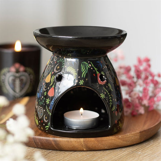 A black ceramic oil burner with floral and heart motifs holds a lit tealight, ideal for wax melts. It rests on a wooden tray with pink and white flowers close by, while a black candle glows gently in the background.