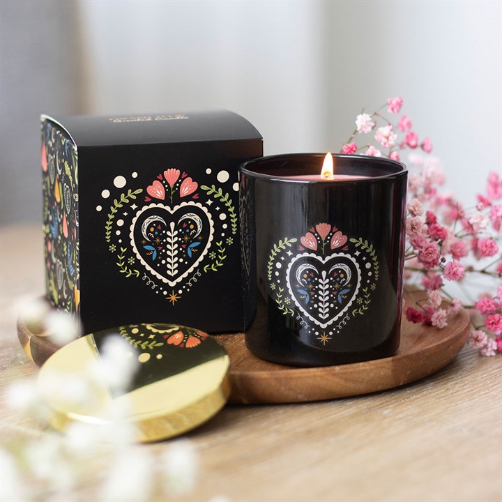 A lit French Tulip scented candle in a glass jar with a colourful, folk-style heart pattern rests on a wooden tray next to its box and gold lid, surrounded by pink and white flowers—a luxurious home fragrance accent.