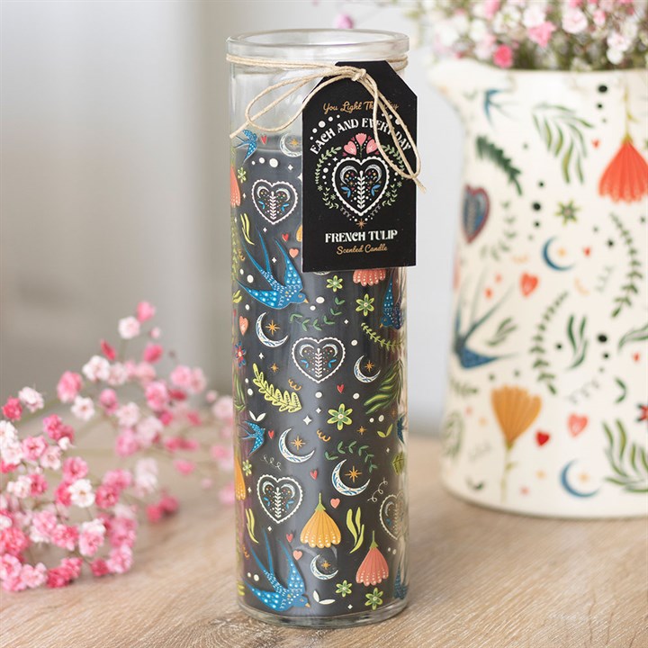 A tall, scented glass candle adorned with colourful floral and heart patterns, labelled “French Tulip.” It rests on a wooden surface next to pink flowers and a white floral vase in the background.