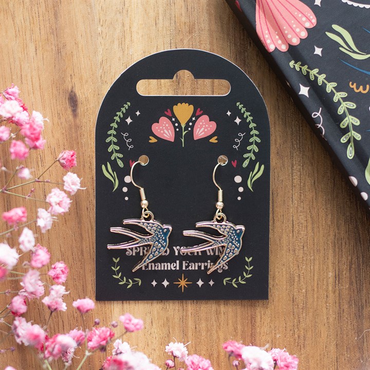 A pair of enamel bird earrings with gold hooks rest on a decorative black card adorned with floral motifs; pink babys breath and an unidentified floral-patterned notebook lie nearby on a wooden surface.