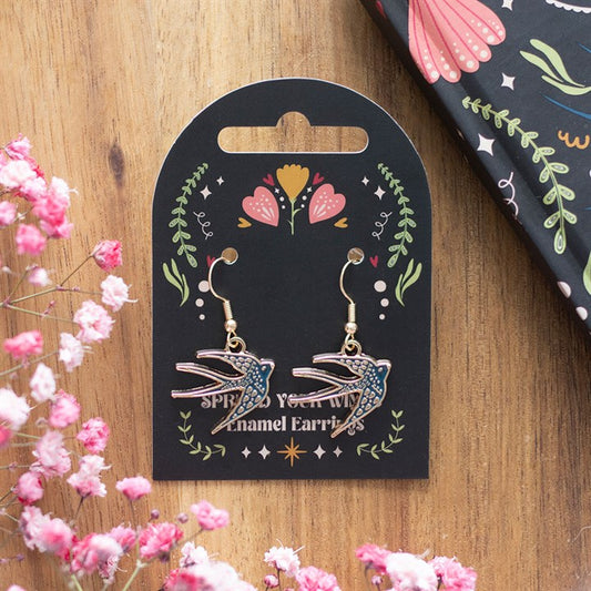 A pair of enamel bird earrings with gold hooks rest on a decorative black card adorned with floral motifs; pink babys breath and an unidentified floral-patterned notebook lie nearby on a wooden surface.