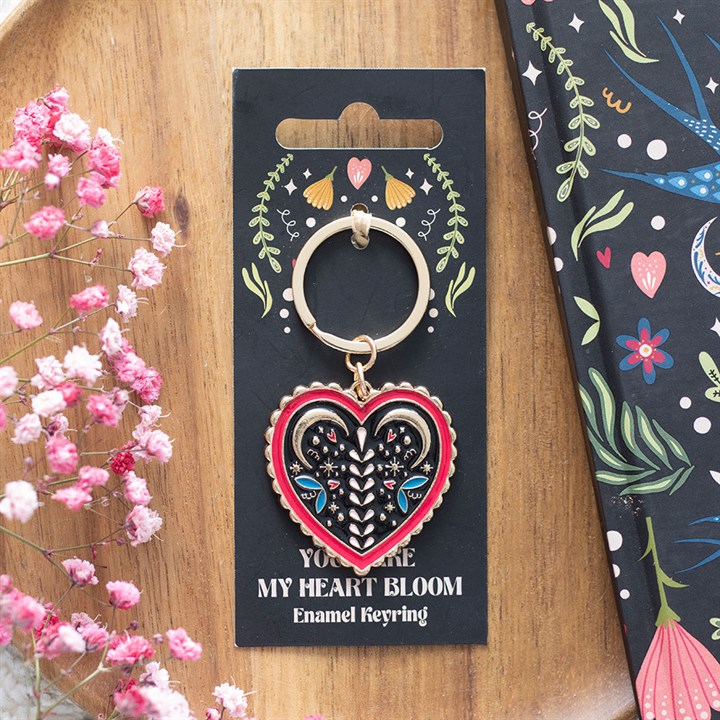 A heart-shaped folk keyring with enamel floral and eye designs rests on a black card reading “YOU MAKE MY HEART BLOOM”, surrounded by pink flowers and a vibrant, floral-patterned notebook.