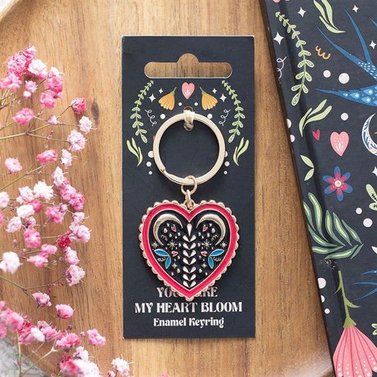 A heart-shaped folk keyring with enamel floral and eye designs rests on a black card reading “YOU MAKE MY HEART BLOOM”, surrounded by pink flowers and a vibrant, floral-patterned notebook.