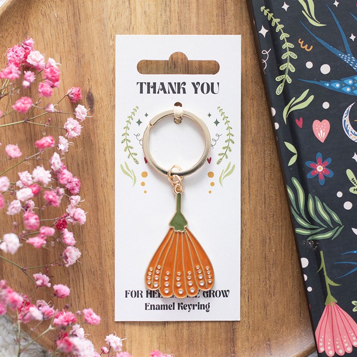 A floral enamel keyring, shaped like an orange flower petal, hangs on a Thank You card with leafy designs—a delightful teacher appreciation gift placed next to pink flowers and a black notebook with colourful floral patterns.